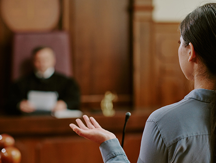 A woman speaks in a courtroom, facing a judge seated at a wooden bench. The scene conveys a serious and formal tone.