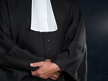 Person wearing a black legal robe and white court tabs, hands clasped in front.
