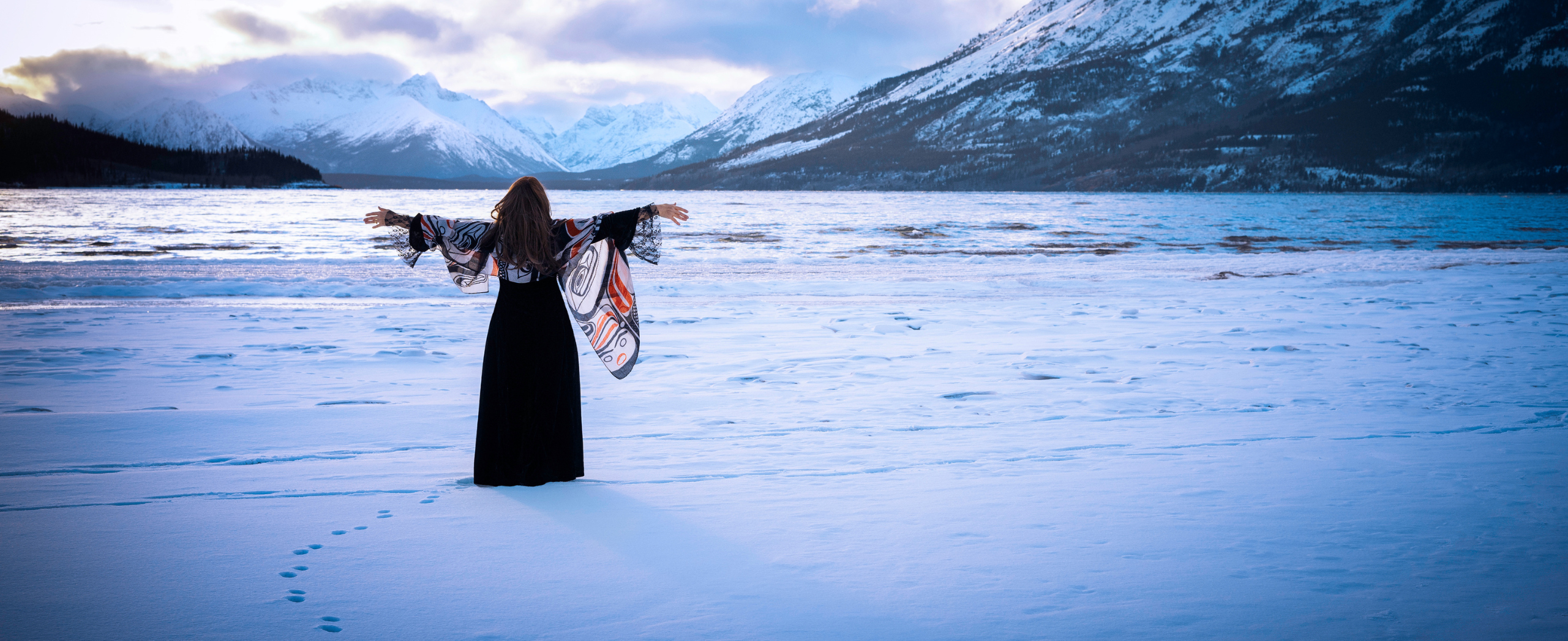 An Indigenous woman stands in the snow near a mountain lake