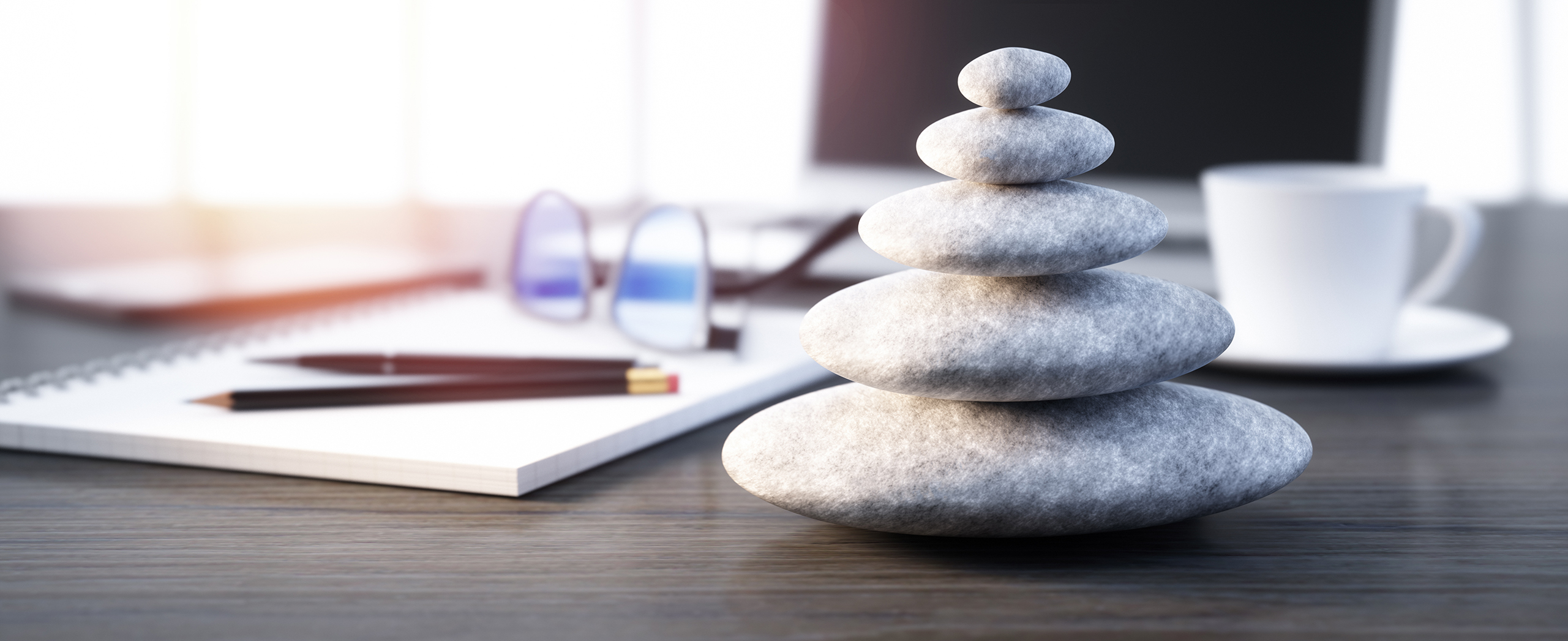 A stack of smooth gray stones sits on a desk, symbolizing balance. Nearby are glasses, a notebook, and a cup, creating a calm, organized workspace.